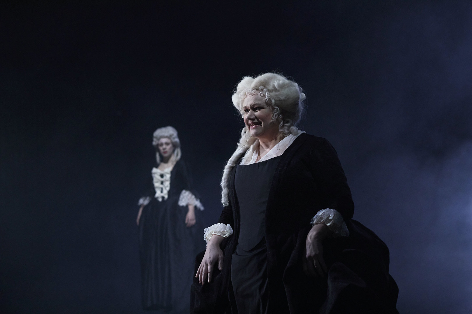 Forestillingsbilde av to kvinnelige skuespillere i barokke kostymer mot mørk bakgrunn