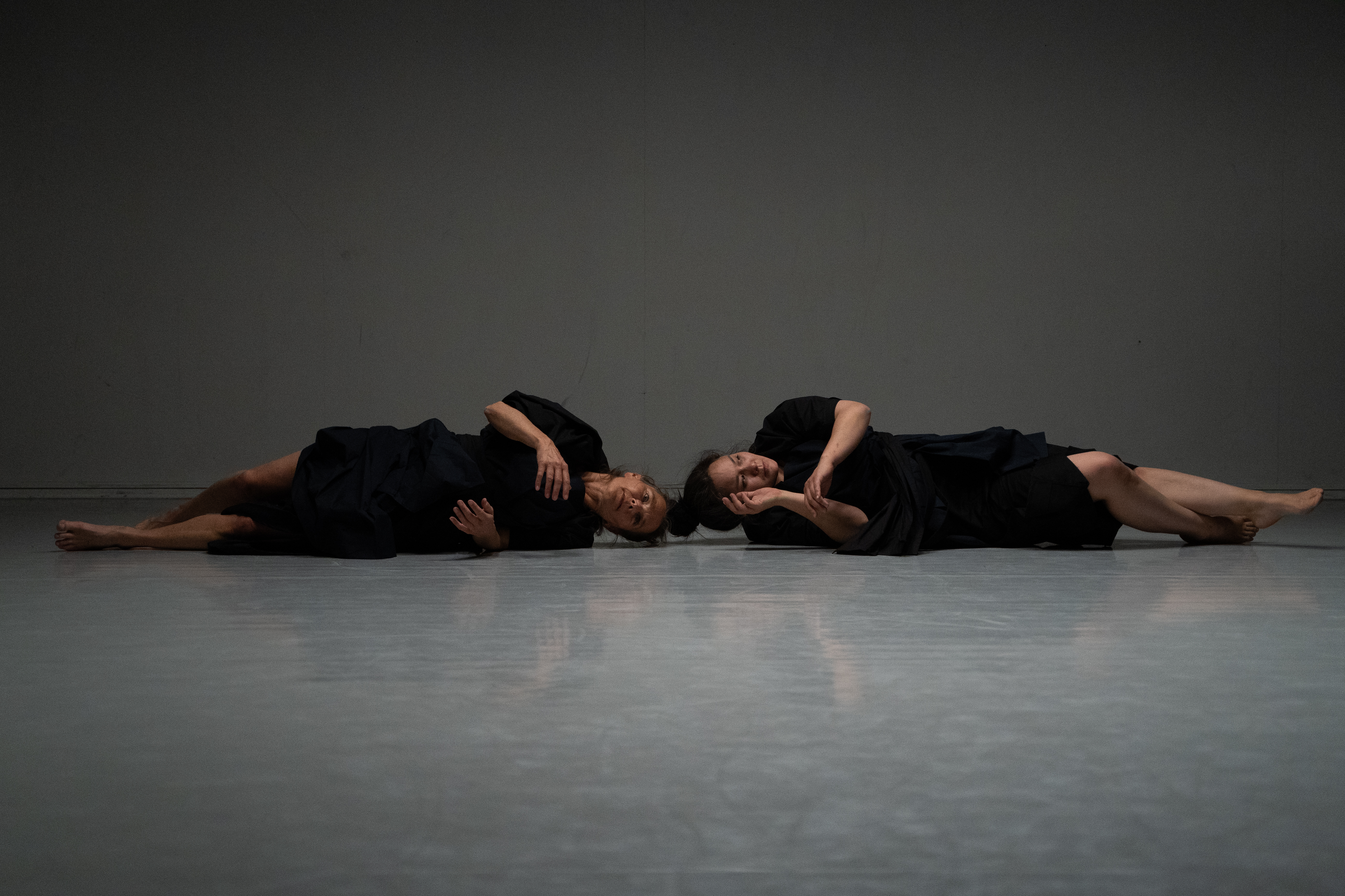 Scenebilde: To kvinner ligger på langs scenen med hode mot hode i sorte klær, slik at hodene danner midten av bildet og føttene i hver sin kant